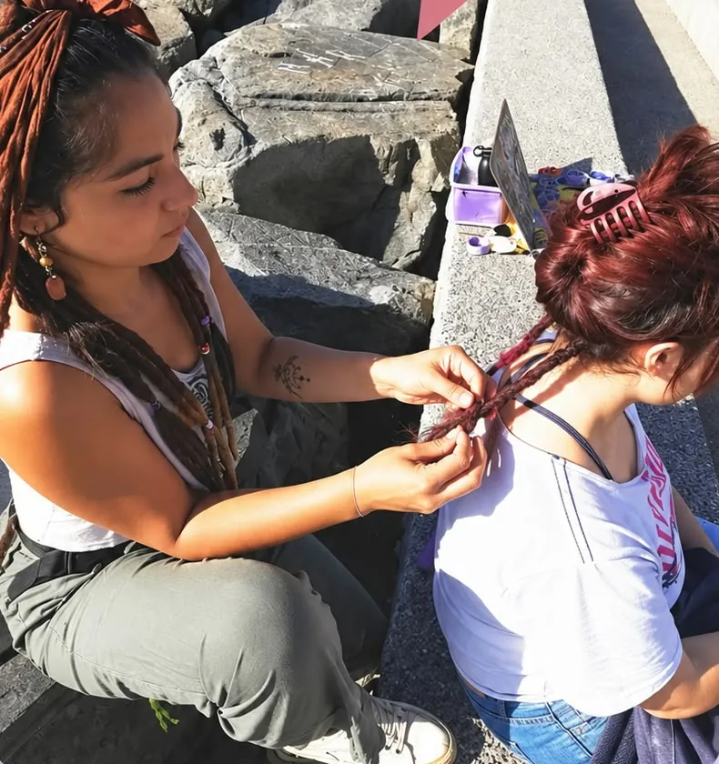 Nae tejiendo dreads a una clienta sentada en un muelle junto al mar